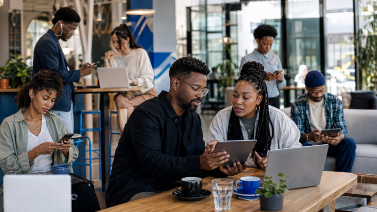 A diverse group of adults uses phones, tablets and laptops while discussing digital content in a modern café.